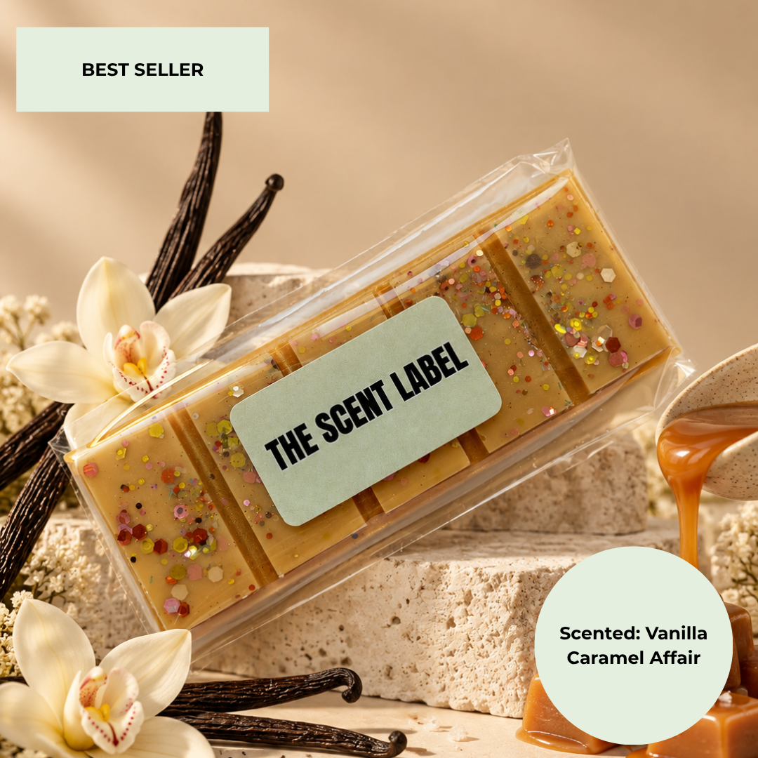 A clear plastic package containing four rectangular bars of tan wax or soap with embedded multicolor glitter, labeled 'THE SCENT LABEL.' The package is styled with vanilla beans, white orchids, and caramel drizzle on a warm beige background.