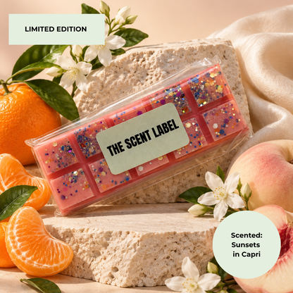 A clear plastic clamshell package containing pink wax melts with blue and gold glitter, labeled 'THE SCENT LABEL,' styled on a stone slab with citrus, peaches, white flowers, and draped fabric. Text overlays read 'LIMITED EDITION' and 'Scented: Sunsets in Capri.