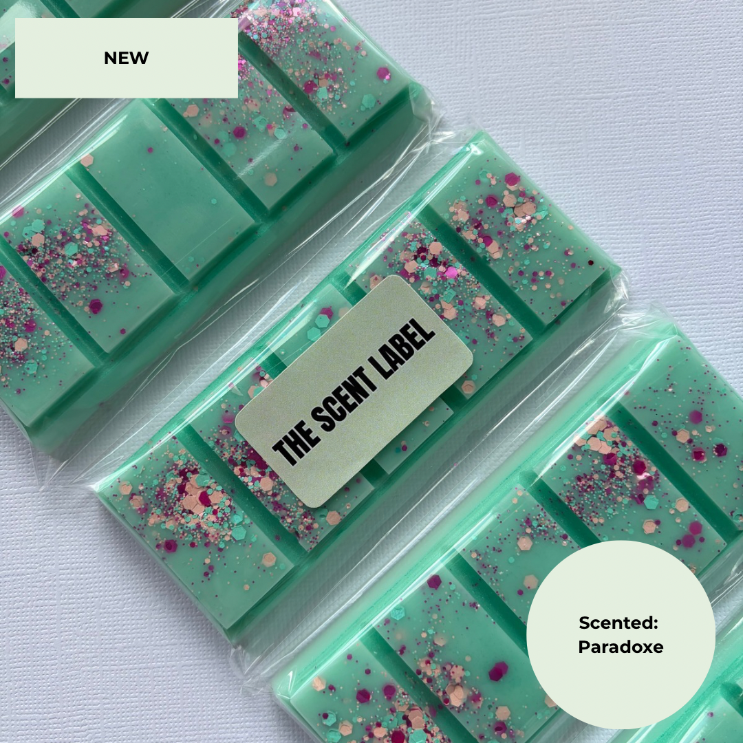 Three packaged mint green wax melts bars with pink and gold glitter, arranged diagonally on a white textured fabric background. A central label reads 'THE SCENT LABEL' and the image includes 'NEW' and 'Scented: Paradox' text overlays.
