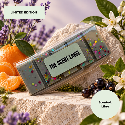 A clear rectangular plastic box containing a gray rectangular product with multicolored confetti, labeled 'THE SCENT LABEL,' styled on a stone surface with citrus, flowers, and berries. Text badges read 'LIMITED EDITION' and 'Scented: Libre'.