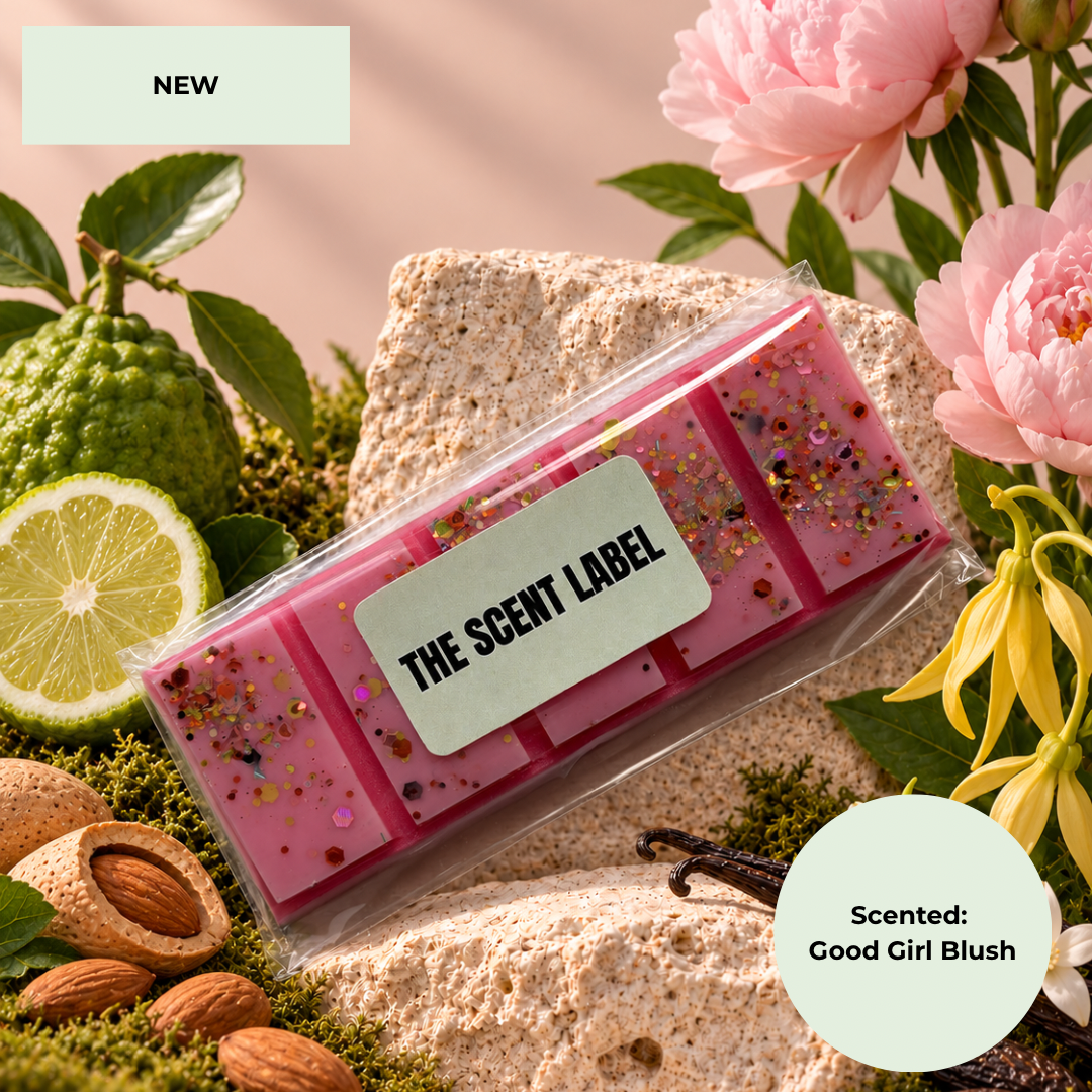 A packaged pink wax melt or scented bar product labeled 'THE SCENT LABEL' lies on a stone surface surrounded by flowers, a lime half, almonds, moss, and vanilla bean. A pale green badge says 'NEW' and another says 'Scented: Good Girl Blush'.