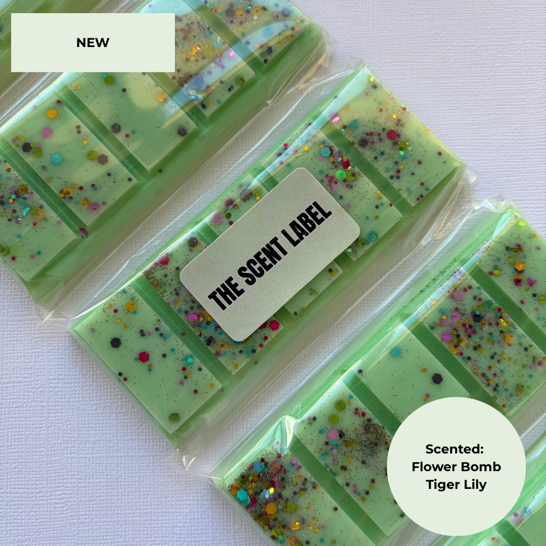 Three packaged green rectangular wax melts with multicolored glitter and flowerbomb inclusions, arranged diagonally on a white textured background. A white label reads 'THE SCENT LABEL,' and text circles say 'NEW' and 'Scented: Flower Bomb Tiger Lily'.