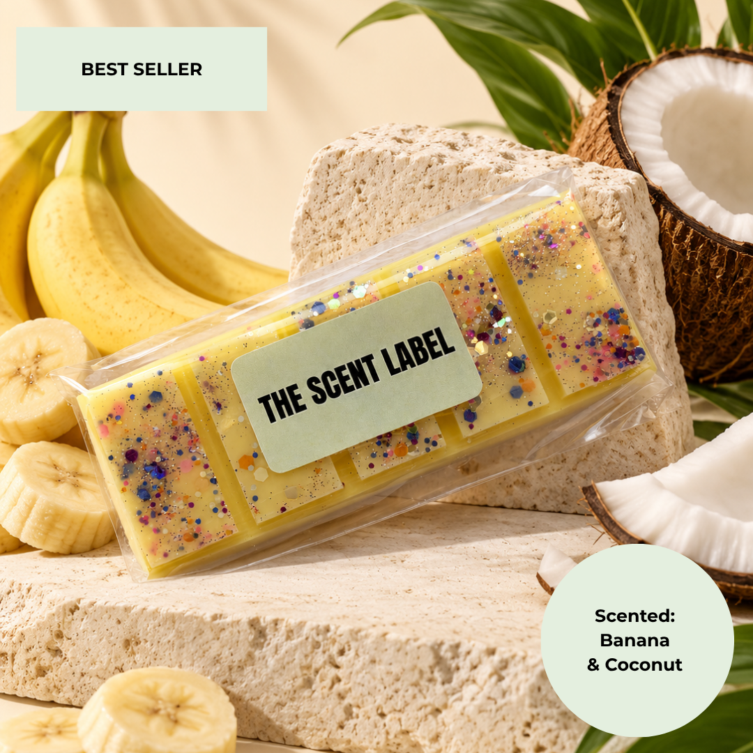 A packaged wax melt bar set from The Scent Label, styled with fresh bananas, sliced bananas, a coconut half, and a textured stone block. The wax melts are yellow with multicolor glitter.