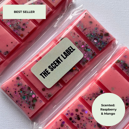 Three pink scented wax melts in clear plastic packaging, decorated with multicolor glitter. A cream-colored label on the center package reads 'THE SCENT LABEL'.