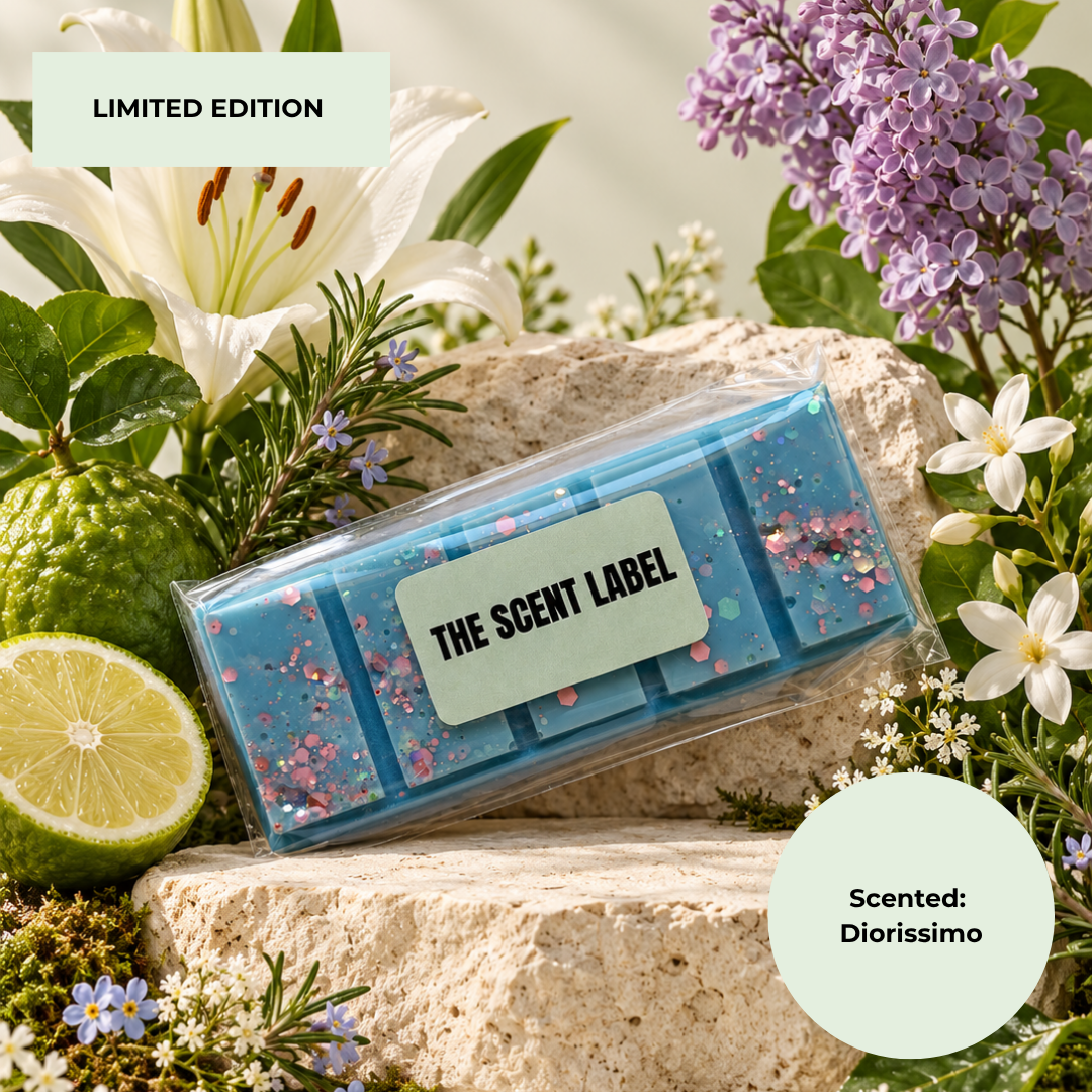 A blue scented wax melt bar in a clear plastic sleeve with a light green label reading 'THE SCENT LABEL', styled on a stone surrounded by flowers, a lime, and greenery. Text overlays read 'LIMITED EDITION' and 'Scented: Diorissimo'.
