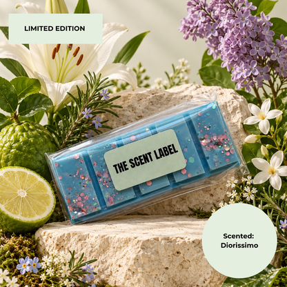 A blue scented wax melt bar in a clear plastic sleeve with a light green label reading 'THE SCENT LABEL', styled on a stone surrounded by flowers, a lime, and greenery. Text overlays read 'LIMITED EDITION' and 'Scented: Diorissimo'.