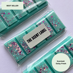 Three packaged teal wax melts or scented bars with pink and blue glitter, arranged diagonally on a light lavender background. A central label reads 'THE SCENT LABEL,' with 'BEST SELLER' in the top-left corner and 'Scented: Daisy Fresh' in a bottom-right circle.