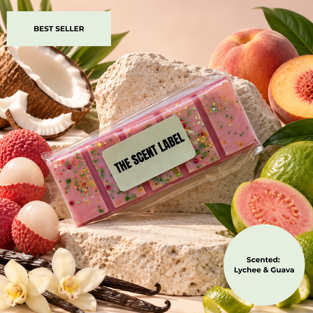 A pink scented wax melt bar with colorful glitter flakes and a light green label reading 'THE SCENT LABEL' is displayed on a stone pedestal. The product is styled with its scent ingredients, including lychee fruits, guava, vanilla beans, a coconut, and white flowers, on a warm beige background.