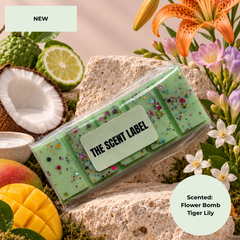 A packaged scented bar product labeled 'THE SCENT LABEL' lies on a stone surface surrounded by tropical props including flowers, a coconut half, lime slices, mango pieces, and moss. A 'NEW' badge appears at top left, and a scent note at bottom right reads 'Scented: Flower Bomb Tiger Lily.