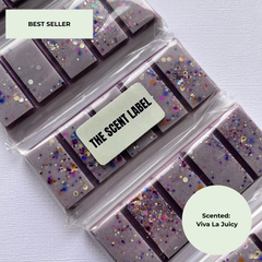 Several clear plastic bags of rectangular lavender-colored bars with multicolor glitter and foil pieces, arranged diagonally on a light fabric background. A central label reads “THE SCENT LABEL,” with a “BEST SELLER” badge in the top left and a circular “Scented: Viva La Juicy” badge in the bottom right.