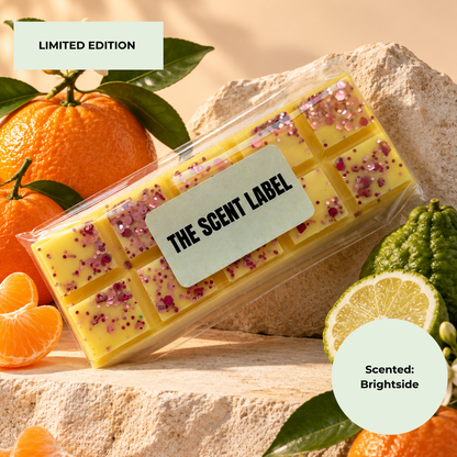 A clear plastic package containing yellow rectangular wax melts  with pink glitter and flower pieces, labeled 'THE SCENT LABEL,' styled with oranges, a kumquat, and a lime slice on a stone surface. Text overlays read 'LIMITED EDITION' and 'Scented: Brightside.