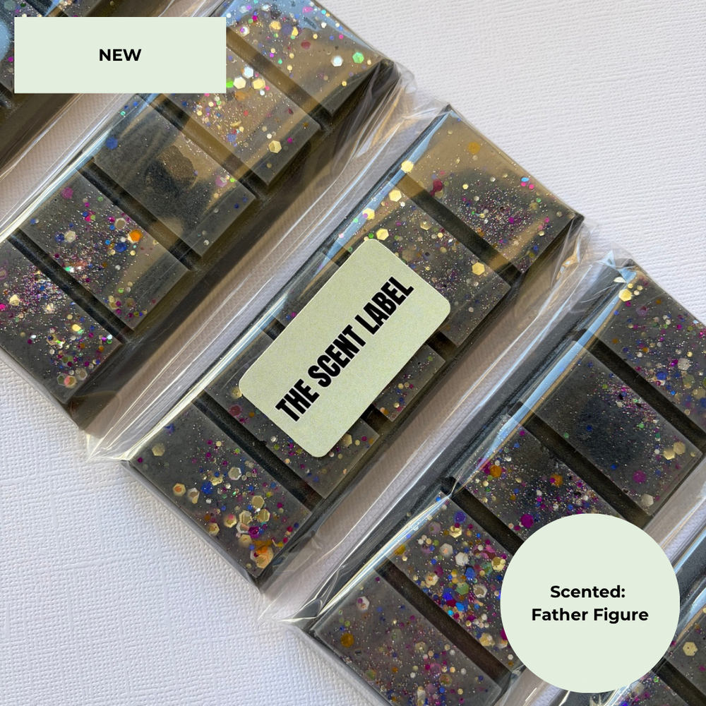 Several clear plastic-wrapped rectangular wax melts or soap bars with embedded multicolor glitter are arranged on a white textured surface. A cream label on one package reads “THE SCENT LABEL,” and a cream badge in the corner says “Scented: Father Figure.