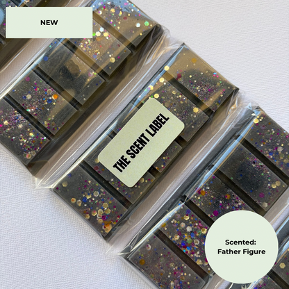 Several clear plastic-wrapped rectangular wax melts or soap bars with embedded multicolor glitter are arranged on a white textured surface. A cream label on one package reads “THE SCENT LABEL,” and a cream badge in the corner says “Scented: Father Figure.