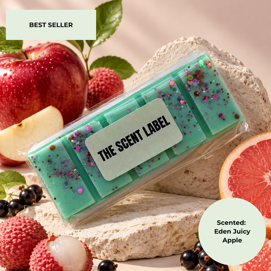 A packaged mint-green scented bar soap or wax melt labeled 'THE SCENT LABEL' rests on a stone slab surrounded by fruit props including a red apple, lychee, blackberries, and a grapefruit. Text badges read 'BEST SELLER' and 'Scented: Eden Juicy Apple'.