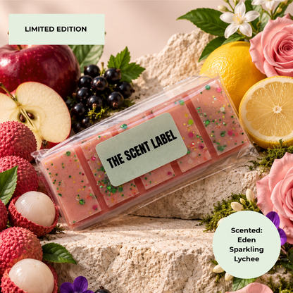 A packaged pink scented wax melt bar set labeled 'THE SCENT LABEL' surrounded by fruit and flowers, including lychee, apples, grapes, lemons, and roses, with text reading 'LIMITED EDITION' and 'Scented: Eden Sparkling Lychee'.