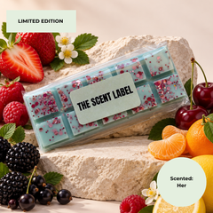 A clear plastic package containing mint-green scented bars with pink floral inclusions and sprinkles, labeled 'THE SCENT LABEL', styled on a stone surface with strawberries, cherries, blackberries, orange slices, and lemon around it. Text badges read 'LIMITED EDITION' and 'Scented: Her'.