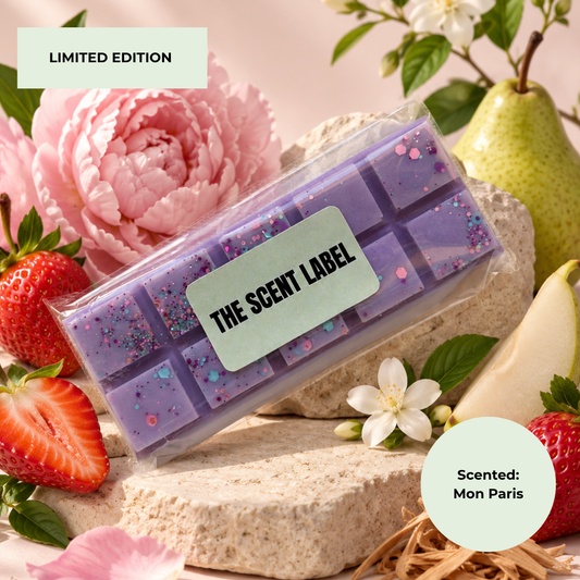 A packaged lavender wax melt bar set with glitter, labeled 'THE SCENT LABEL', styled with strawberries, a pink flower, a pear, and stone blocks. Text badges read 'LIMITED EDITION' and 'Scented: Mon Paris'.
