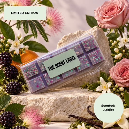 A clear plastic clamshell package containing rectangular purple scented bars with glitter and flower inclusions, placed on a stone surface surrounded by pink roses, blackberries, white flowers, green leaves, and vanilla beans. Text overlays read 'LIMITED EDITION' and 'Scented: Addict'; the label on the package reads 'THE SCENT LABEL'.